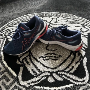 Blå Asics sneakers med röda detaljer - Snygga blå sneakers från Asics med röda detaljer och vit sula. Ovandelen är i mesh för bra andningsförmåga och skorna har klassisk snörning. Perfekta för dig som gillar sportig stil och vill ha bekväma skor till vardags.