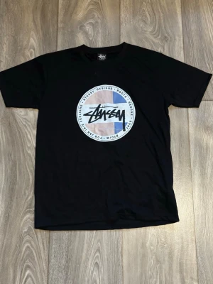 Svart Stussy t-shirt med tryck - Säljer en svart t-shirt från Stussy med stort runt tryck på bröstet i blått, beige och vitt. T-shirten har klassisk passform och korta ärmar. Perfekt för dig som gillar streetwear och vill ha något med en ikonisk logga.