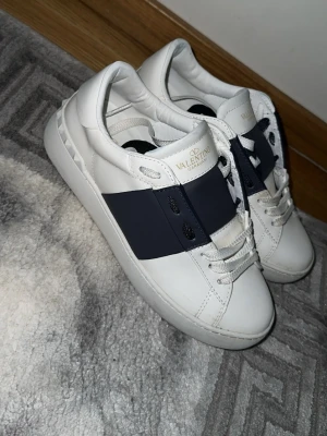 Valentino vita sneakers med blå detalj - 9/10 skick använt endast 2-3 gånger.Snygga vita sneakers från Valentino Garavani med bred mörkblå remsa över sidan och coola nitar på hälen. Skorna har vita skosnören och är tillverkade i slätt läder. Perfekta för dig som vill ha en stilren men ändå unik look.