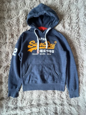 Mörkblå Superdry hoodie med tryck - Snygg mörkblå hoodie från Superdry med stort gult och vitt tryck på bröstet och siffran 23 på ärmen. Klassisk känguruficka framtill och vita dragsnören i huvan. Tillverkad i mjukt bomullsmaterial, perfekt för en avslappnad streetwear-look.