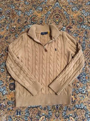 Beige kabelstickad tröja Polo Ralph Lauren - Kabelstickad beige tröja från Polo Ralph Lauren med halv dragkedja och hög krage. Tröjan har långa ärmar, ribbade muddar och nederkant samt den klassiska broderade loggan på bröstet. Perfekt för lager på lager och snygg till jeans. Den sitter bra på På S och M