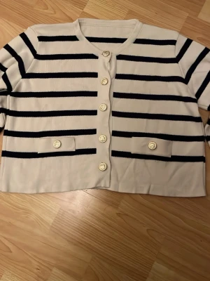 Randig kofta med guldknappar - Snygg beige kofta med svarta horisontella ränder och dekorativa guldknappar framtill. Koftan har rund halsringning, två fejkfickor med knappdetaljer och en croppad passform. Perfekt för en trendig och stilren look.