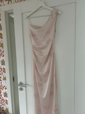 Ljusrosa långklänning med blommor - Superfin ljusrosa balklänning med blommiga mönster från Adoore. Klänningen har draperad detalj framtill och är tillverkad i ett glansigt, mjukt material som ger en lyxig känsla. Perfekt för speciella tillfällen. Endast använd en gång och säljer då den tyvärr är för liten. Köpt för 1600.
