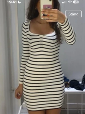 Vit och svart randig kortklänning - Säljer en vit kortklänning med svarta horisontella ränder. Klänningen har lång ärm och en rundad urringning. Materialet ser ut att vara mjukt och stretchigt, perfekt för en trendig och bekväm look.