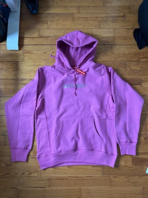 rosa Supreme hoodie med huva - Supreme hoodie i en snygg rosa färg med klassisk huva och magficka. Loggan sitter broderad på bröstet och dragsko i halsen har Supreme-tag. Tillverkad i mjuk bomullsblandning som känns skön mot huden. Perfekt för dig som vill sticka ut med streetwear-vibe. !!!ser mer rosa ut i verkligheten!!!’