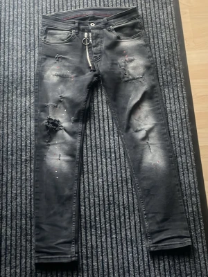 Svarta slim fit jeans - Säljer ett par svarta slim fit jeans med slitningar och trasiga detaljer framtill. Jeansen har en silverfärgad dragkedja med ringdetalj och små färgstänk. Klassisk femficksmodell och raka bakfickor. Materialet är troligtvis bomull med stretch.