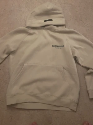 Beige hoodie Essentials Fear of God - Snygg beige hoodie från Essentials Fear of God med svart logga på bröstet och ärmen. Hoodien har stor huva, magficka och ribbade muddar. Tillverkad i mjukt material som känns riktigt skönt mot huden. Perfekt för dig som gillar streetwear och enkel stil.