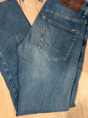 Blå slim fit jeans från G-Star RAW - Snygga blå slim fit jeans från G-Star RAW med klassisk femficksdesign och läderpatch bak i midjan. Jeansen har en lätt tvättad look och smal passform som sitter tajt längs benen. Tillverkade i slitstark denim med subtila detaljer.