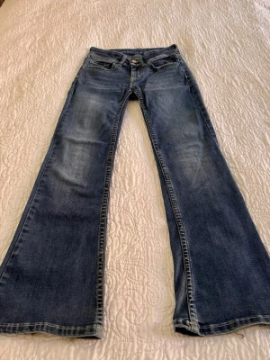 Blå bootcut jeans med slitningar - Snygga blå bootcut jeans med lätt slitning och utsvängda ben. Klassisk femficksmodell med dragkedja och knapp framtill. Jeansen har en tvättad look och råa kanter nertill för en cool vibe. Perfekta för dig som gillar retrostil och avslappnad känsla.
