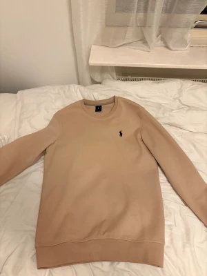 Beige sweatshirt från Ralph Lauren - Snygg beige sweatshirt från Ralph Lauren med klassisk rund halsringning och den ikoniska svarta logotypen broderad på bröstet. Tröjan har ribbade muddar vid ärmslut och nederkant, perfekt för en clean och stilren look.