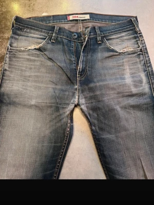 Levi's 504 blå slitna jeans - Säljer ett par blå Levi's 504 jeans med snyggt slitna detaljer vid fickorna och klassisk tvättad look. Jeansen har raka ben, fem fickor och orange sömmar. Perfekta för en avslappnad och trendig stil.