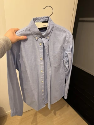 Ljusblå skjorta - Klassisk ljusblå skjorta från Jack and Jones med button-down krage och vita knappar. Skjortan har en bröstficka och lång ärm, tillverkad i mjuk bomull. Perfekt för en clean och stilren look.