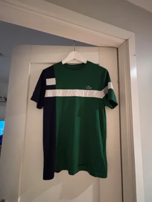 Grön och marinblå Lacoste t-shirt - Säljer en cool t-shirt från Lacoste i grönt och marinblått med vita detaljer. T-shirten har ett sportigt snitt, rund hals och logotyp på bröstet samt på ärmen. Materialet är mjukt och bekvämt, perfekt för träning eller chill. Passar dig som gillar stilren design.