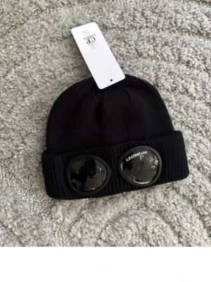 Svart mössa med glasögondetalj C.P. Company - Säljer en svart ribbstickad mössa från C.P. Company med ikoniska glasögondetaljer på uppviket. Mössan är tillverkad i mjukt material och har en modern, streetinspirerad look. Perfekt accessoar för dig som gillar trendiga detaljer.