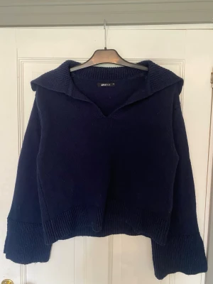 Mörkblå stickad tröja Gina tricot - Mysig mörkblå stickad tröja från Gina tricot med bred krage och v-ringning. Tröjan har långa, vida ärmar och ribbstickade muddar vid ärmslut och nederkant. Perfekt för kyliga dagar och enkel att styla till jeans eller kjol.