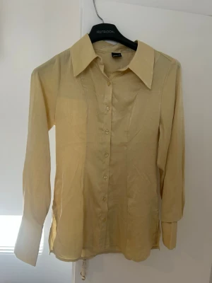 Gul skjorta i sidenlook - Stilren gul skjorta från Gina tricot med lång ärm och klassisk krage. Skjortan har knappar framtill och är gjord i ett glansigt material som ger en lyxig sidenkänsla. Perfekt för dig som gillar en clean och tidlös stil.