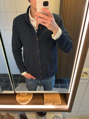 Mörkblå kofta med träknappar H&M - Mysig mörkblå kofta från H&M med ribbad krage och muddar. Koftan har träfärgade knappar framtill och två fickor. Perfekt att slänga över en skjorta för en avslappnad men snygg look.