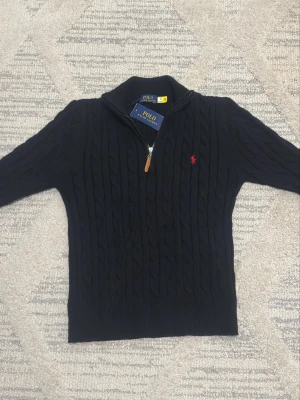 Svart kabelstickad tröja Polo Ralph Lauren - Kabelstickad svart tröja från Polo Ralph Lauren med halv dragkedja och klassisk röd logga broderad på bröstet. Tröjan har hög krage och ribbade muddar. Perfekt för kalla dagar och snygg till jeans eller chinos.