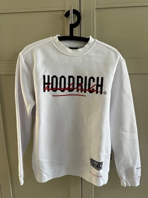 Vit sweatshirt från Hoodrich - Säljer en vit sweatshirt från Hoodrich med svart och rött tryck på både fram- och baksidan. Tröjan har rund halsringning, långa ärmar och detaljerade broderier samt patch nertill. Perfekt för dig som gillar streetwear och vill sticka ut.