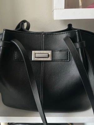 Svart bucket bag med silverdetalj - Stilren svart handväska i bucket-modell med långa axelband och en snygg silverfärgad rektangulär detalj framtill. Väskan har en mjuk, rundad form och är tillverkad i slätt konstläder. Perfekt för dig som gillar minimalistisk och modern design🤍💞