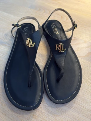 Svarta lädersandaler från Lauren Ralph Lauren - Stilrena svarta sandaler i läder från Lauren Ralph Lauren med guldfärgade RLL-detaljer framtill. Klassisk t-rem design med justerbar rem runt hälen och diskreta sömmar längs sulan. Perfekta för en trendig och avslappnad look.
