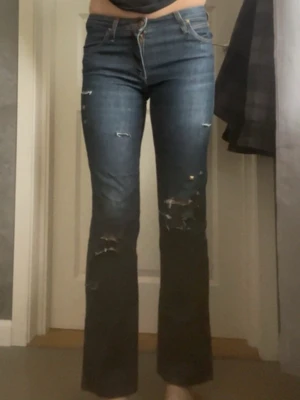 Blå bootcut jeans med nitar - Säljer ett par blå bootcut jeans med slitningar och råa kanter. Jeansen har coola guldfärgade nitar på ena benet och klassisk femficksmodell. Perfekta för dig som gillar en edgy och trendig stil.