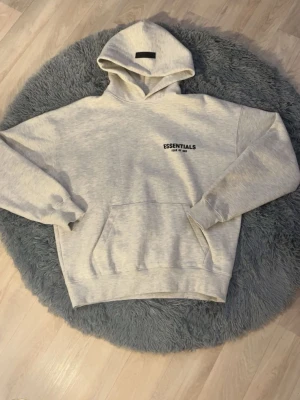 Grå Essentials Fear of God hoodie - Säljer en ljusgrå hoodie från Essentials Fear of God med svart tryck fram och stort tryck på ryggen. Hoodien har huva, känguruficka och ribbade muddar. Perfekt oversized streetwear-look och riktigt skön bomullskänsla.