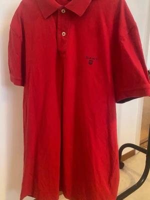 Röd pikétröja från Gant - Klassisk röd pikétröja från Gant med korta ärmar, krage och två knappar framtill. Tröjan har en broderad Gant-logga på bröstet och är tillverkad i mjuk bomull. Perfekt för en avslappnad men ändå stilren look.