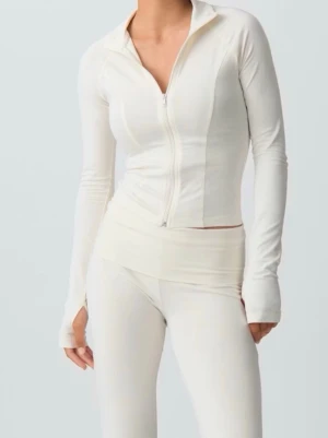 Vit loungewear-set med dragkedja - Superfräscht vitt loungewear-set med långärmad ziptröja och matchande byxor. Tröjan har hög krage, dragkedja framtill och tumhål i ärmsluten. Setet är figurnära och gjort i mjukt, stretchigt material som känns skönt mot huden.