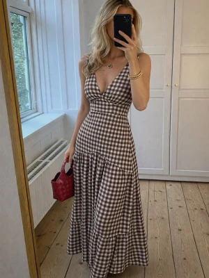 Brun och vit rutig långklänning - Snygg långklänning med brunt och vitt rutigt mönster och djup v-ringning. Klänningen är ärmlös och har en figurnära passform upptill med volangdetalj nedtill. Perfekt för sommardagar och picknickar.