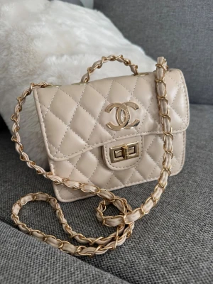 Beige quiltad  handväska - Supersnygg beige quiltad handväska med klassisk CC-logga i guld framtill. Väskan har en kedjerem med läderdetaljer och ett elegant vridlås. Insidan är rymlig med en innerficka med dragkedja. Perfekt accessoar för att lyfta vilken outfit som helst.