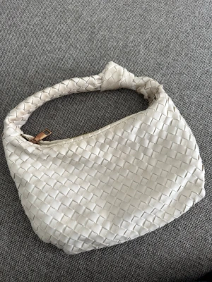 Vit flätad axelväska - Snygg vit handväska med flätad design och mjukt material. Väskan har ett kort axelband med knutdetalj och stängs med dragkedja i guld. Perfekt storlek för det viktigaste och ger en trendig touch till din outfit.