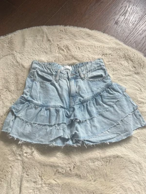 Ljusblå volangkjol i jeans - Supersöt kort jeanskjol i ljusblå denim med volanger och råa kanter. Kjolen har klassiska fickor fram och bak, bälteshällor och dragkedja framtill. Perfekt för dig som gillar en avslappnad men trendig stil.