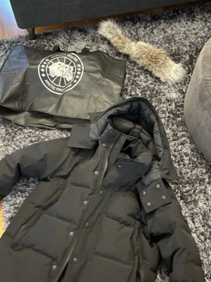 Svart dunjacka från Canada Goose - Säljer en svart dunjacka från Canada Goose med avtagbar huva och pälskant. Jackan har stora fickor framtill, dragkedja och knappar samt klassisk logga på ärmen. Perfekt för kalla vinterdagar och riktigt varm. Priset kan diskuteras. Den har endast används 3 gånger. Så gott som ny 