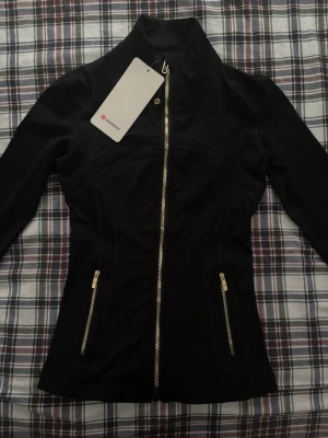 Svart träningsjacka från Lululemon - Snygg svart träningsjacka från Lululemon med hel dragkedja framtill och två fickor med dragkedja. Figurnära passform och hög krage. Perfekt för gymmet eller löprundan. Tillverkad i stretchigt funktionsmaterial.