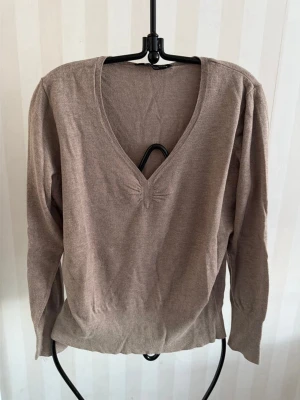 Beige stickad tröja med v-ringning - En enkel och stilren beige stickad tröja med djup v-ringning och diskret detalj vid halsen. Tröjan har långa ärmar och ribbade muddar vid ärmslut och nederkant. Perfekt för lager på lager och passar till det mesta.