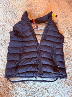 Mörkblå dunväst från Parajumpers - Snygg mörkblå dunväst från Parajumpers med quiltad design och två dragkedjeförsedda fickor framtill. Västen har hög krage och ikonisk gul detalj vid halsen. Perfekt för lager-på-lager och håller dig varm under kyliga dagar. Den är även i nästan oanvänt skick.