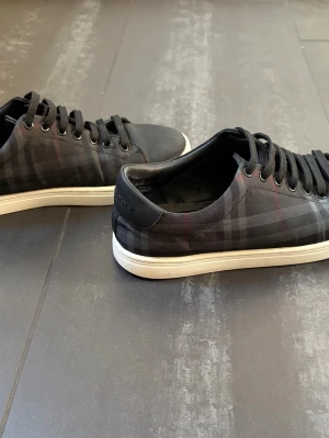 Burberry skor - Snygga svarta sneakers från Burberry med klassiskt rutmönster i grått och diskreta röda detaljer. Skorna har vita sulor, svarta skosnören och metallöljetter. Tillverkade i textil och läderdetaljer vid hälen där Burberry-loggan syns. Perfekta för en trendig streetstyle-look.