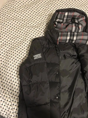Svart pufferjacka från Burberry - Säljer en svart pufferjacka från Burberry med huva och klassiskt rutigt foder i grått, vitt och rött. Jackan har en patch med Burberry London England på ärmen och dragkedja framtill. Perfekt för kalla vinterdagar och riktigt snygg streetstil.Kan också användas som väst under våren/ sommaren 