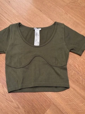 Olivgrön ribbad croppad topp - Trendig olivgrön croppad topp från Bershka med ribbad struktur och markerad söm under bysten. Toppen har korta ärmar och rund halsringning, perfekt för en cool och avslappnad stil.