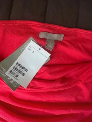 Röd bandeau tubtopp från H&M - Snygg röd bandeau tubtopp från H&M i mjuk bomullsmix. Toppen är axelbandslös och har rynkade sidor för en cool och bekväm passform. Perfekt att styla med jeans eller kjol för en trendig look. Korall färgad