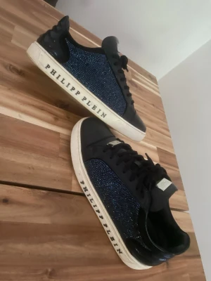 Philipp Plein mörk blå sneakers med blå glitter - Säljer ett par svarta Philipp Plein sneakers med blå glittriga detaljer på sidorna och vit sula med logotyp. Skorna har svarta skosnören, metallplatta på plösen och är tillverkade i läder och textil. Skrona är köpt för 10000 kr ungefär 
