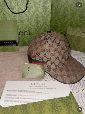 Gucci keps med GG-mönster - Snygg keps från Gucci i beige och brunt med klassiskt GG-monogram över hela kepsen. Har en grön och röd rand på sidan som signaturdetalj. Bågen är böjd och materialet är canvas. Perfekt för dig som vill ha en lyxig accessoar med streetkänsla. Alla tillbehör följs med förutom kvitton 