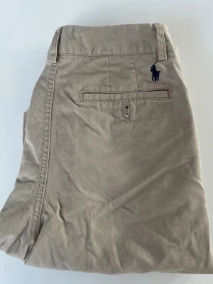 Beige shorts från Polo Ralph Lauren - Klassiska beige shorts från Polo Ralph Lauren med broderad logga ovanför bakfickan. Modellen har raka ben, bälteshällor och knappgylf. Tillverkade i mjuk bomull för en avslappnad och stilren look. Vet inte vad storleken är den passar 10-12års åldern.