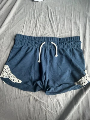 Blå shorts med spetsdetaljer - Mörkblå shorts med vita spetsdetaljer på sidorna och snörning i midjan. Mjukt material och elastisk midja för skön passform. Perfekta för sommardagar eller att chilla hemma.