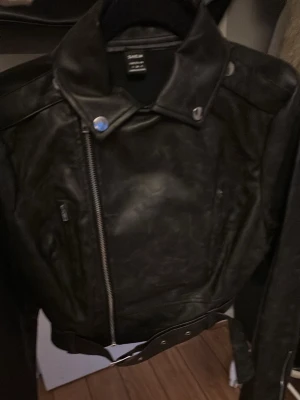 Svart biker skinnjacka från Saint - Snygg svart skinnjacka i klassisk bikerstil från Saint. Jackan har sned dragkedja, bred krage med tryckknappar och bälte i midjan. Två fickor med dragkedja framtill och coola detaljer som ger en edgy look.