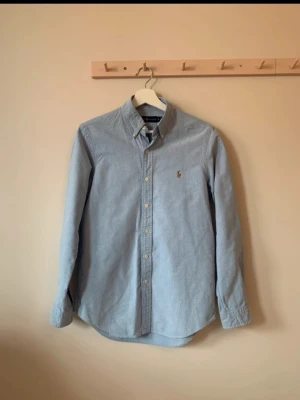 Ljusblå skjorta från Ralph Lauren - Klassisk ljusblå skjorta från Ralph Lauren med button-down krage och broderad logga på bröstet. Skjortan har långa ärmar, knappar framtill och är tillverkad i mjuk bomull. Perfekt för en stilren och tidlös look.