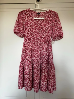 Rosa blommig kortklänning med puffärm - Supersöt rosa kortklänning med vitt blommönster och puffiga kortärmar. Klänningen har markerad midja med knytband i sidan och en luftig kjol. Perfekt för varma dagar och ger en romantisk vibe.