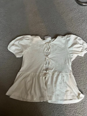 Vit blus med puffärm och knytning - Söt vit blus med korta puffärmar och knytning framtill. Blusen har en lätt utställd nederdel och rund halsringning. Perfekt för en avslappnad och trendig look.