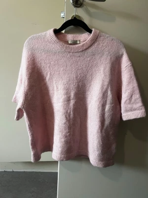 Ljusrosa stickad t-shirttröja - Mysig ljusrosa stickad t-shirttröja från Filippa K med korta ärmar och rund halsringning. Tröjan har en avslappnad passform och ribbade kanter vid hals, ärmslut och nederkant. Perfekt för lager-på-lager och en söt touch till din outfit.